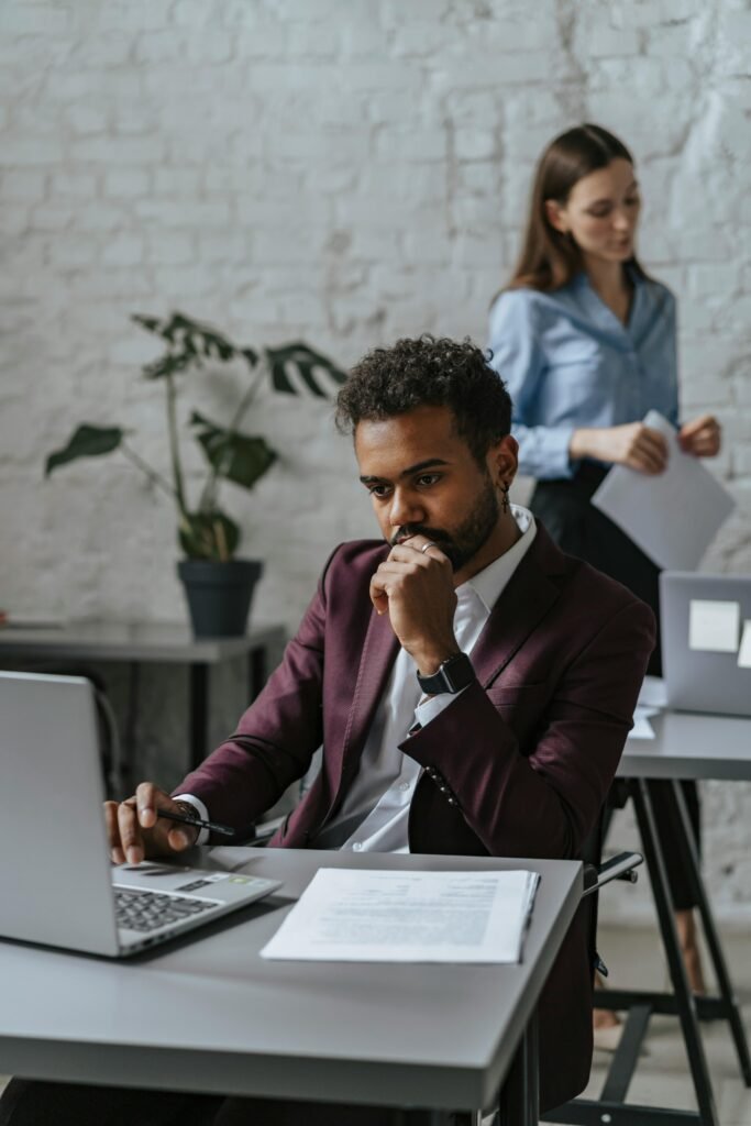 Pensive man using laptop in modern office with female colleague in background.